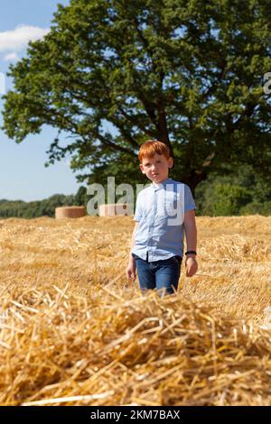 Ein Junge mit roten Haaren läuft auf einem Feld mit drei-Tage- und Strohstapeln aus Weizen, ein Weizenfeld mit einem Jungen während eines Spaziergangs in der Sommersaison, ein Junge von Stockfoto