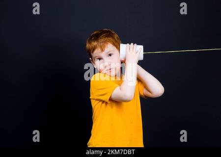 Porträt eines netten Jungen mit roten Haaren, die auf einem einfachen hausgemachten Telefon spricht, spielt der Junge mit anderen Kindern in der Ferne, hält in Kontakt mit einfachen Stockfoto