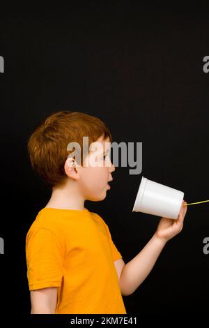 Porträt eines netten Jungen mit roten Haaren, die auf einem einfachen hausgemachten Telefon spricht, spielt der Junge mit anderen Kindern in der Ferne, hält in Kontakt mit einfachen Stockfoto