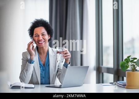 Bild einer reizenden, erwachsenen Unternehmerin, die ein Glas Wasser hält, ein Telefongespräch führt. Stockfoto