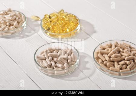 Nahrungsergänzungsmittel in Kapseln in Petrischalen auf weißem Holztisch. omega 3 und verschiedene Vitaminpillen zur Unterstützung des Herz- und Gefäßsystems Stockfoto