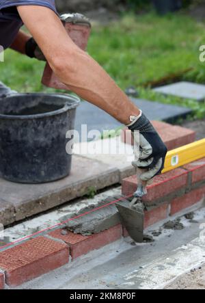 Bauarbeiten im Gange. Männlicher Baumeister, der mit roten Ziegeln arbeitet. Nahaufnahme der Mauerwand. Stockfoto