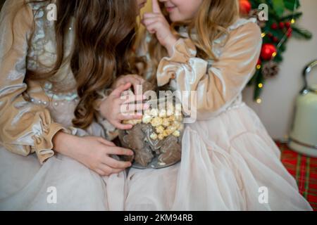 Zwei süße kleine Mädchen in beigen Kleidern sitzen auf dem Tisch und halten Kekse. Neujahrsdekoration. Luxuriöse Neujahrseinrichtung. Stockfoto