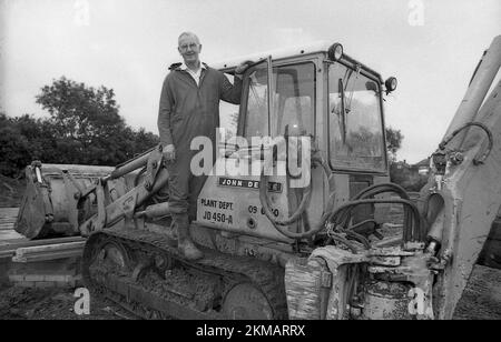 1980er, historisch, draußen auf einer Baustelle, ein einteiliger Overall eines Bauunternehmers, der auf einem John Deere 450-Kettenlader steht, England, Großbritannien. Das 450-Modell wurde 1965 eingeführt und war seit vielen Jahren ein beliebter Bulldozer. Stockfoto