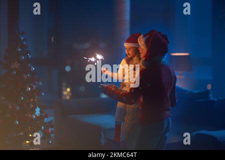 Fröhliche weiße Mutter mit zwei Töchtern, die in weihnachtsmannhüten in der Nähe des Weihnachtsbaums stehen und Funkeln halten. Familie feiert Neujahr zu Hause. Stockfoto