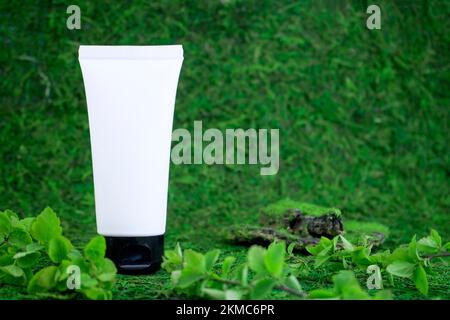 Weiße unbeschriftete kosmetische Flasche, auf einem grünen natürlichen Hintergrund, Gras, Moos mit Ästen von Grün. Umweltfreundliches Kosmetikprodukt. Natürlicher Kosmos Stockfoto