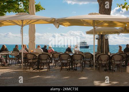 Gardasee Italien, Blick auf die Menschen, die sich an Cafétischen entlang der Uferpromenade in der malerischen Altstadt von Gardasee, Gardasee, Veneto entspannen Stockfoto
