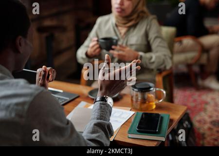 Hände eines jungen afroamerikanischen Geschäftsmannes in grauem Hemd, der Stift hält, während er seine Sichtweise gegenüber einer muslimischen Kollegin erläutert Stockfoto