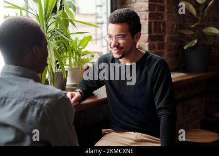 Ein junger lächelnder Geschäftsmann in Freizeitkleidung, der einen afroamerikanischen männlichen Kollegen mit einem Lächeln ansieht, während er über Organisationspunkte spricht Stockfoto