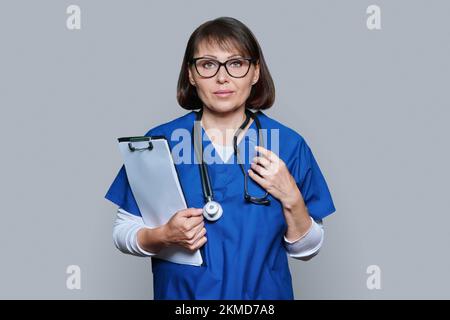 Porträt einer Ärztin mit Clipboard, das auf grauen Hintergrund in die Kamera schaut Stockfoto