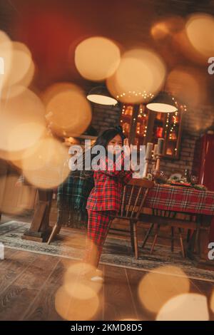 Active japan Kid Girl mit stilvollen langen Brünette Haarmodell zu Hause in Bokeh Girlanden noel posieren. Asiatische kleine Fashionista in rot karierten Pyjama tanzen Stockfoto