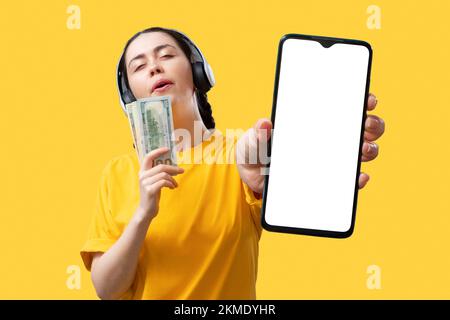 Börse und Einkommen. Die fröhliche junge Weiße mit Kopfhörern zeigt ein Smartphone mit einem Modell-Bildschirm und einen Stapel Dollar Stockfoto