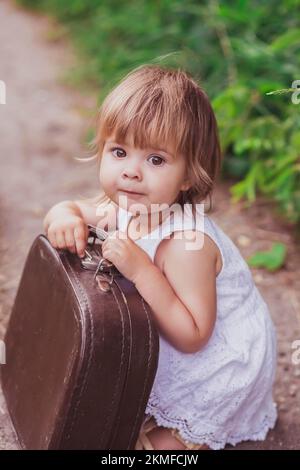 Ein bezauberndes Baby in einem weißen Kleid versucht, einen alten Koffer zu öffnen Stockfoto
