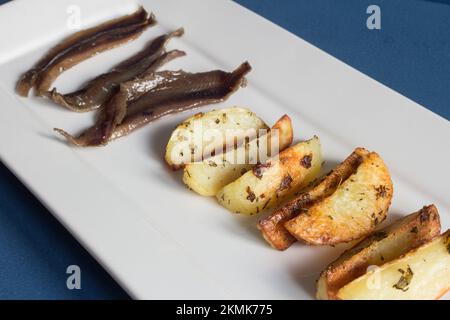 Eine rechteckige weiße Keramikplatte mit gebackenen Rosmarin-Kartoffeln im Vordergrund und Sardellen im Hintergrund. Traditionelle Tapas und Speisen. Stockfoto