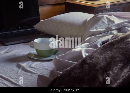 Ein leerer Becher auf einer dreckigen Untertasse neben einem offenen Laptop auf einem ungemachten Schlafzimmerbett mit zerrüttelten Decken. Unordentliches Zimmer. Stockfoto