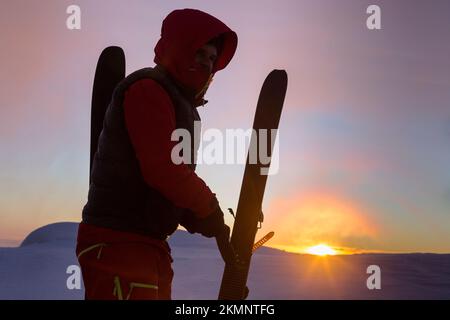 Der Skifahrer zieht die Skier auf dem Hintergrund des orangefarbenen Sonnenaufgangs der Sonnenscheibe in den schneebedeckten Bergen von den Skiern ab Stockfoto