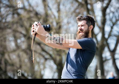 Ein bärtiger Mann, der Selfie auf einer Oldtimer-Kamera macht. Mann mit Bart und Schnurrbart auf fröhlichem Gesicht, Zweige im Hintergrund, Unschärfe. Mann mit langem Bart beschäftigt Stockfoto