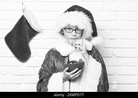 Junge süße weihnachtsmann Junge mit Gläsern in rotem Pullover und Neujahr Hut mit dekorativen weihnachten oder Weihnachten Strumpf oder Stiefel mit Schokolade Hüfte Stockfoto