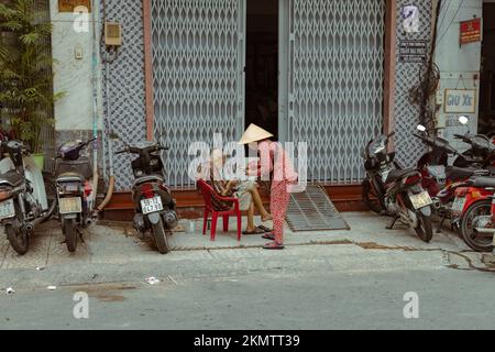 Ho-Chi-Minh-Stadt, Vietnam - 8. November 2022: Eine ältere Frau in traditioneller vietnamesischer Kleidung und ohne Kopfschmuck verkauft Lotteriekarten an einen alten Mann Stockfoto