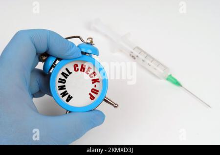 Medizinisches Konzept. Auf dem Tisch befindet sich eine Spritze, ein Wecker in seiner Hand mit einer Inschrift auf der Anzeigetafel - JÄHRLICHE KONTROLLE Stockfoto
