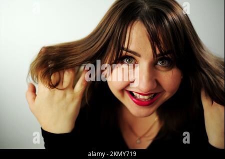 Eine junge Frau lacht auf ihrem Gesicht, ihr Lächeln und ihre Augen leuchten Erwachsene kurvenreiche wunderschöne Frau schaut in den Rahmen Nahgesicht Schwarzes Kleid und roter Lippenstift Sie hat braune Haare Stockfoto