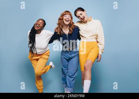 Drei Freundinnen lachen glücklich, während sie sich umarmen. Eine Gruppe fröhlicher junger Frauen, die Spaß haben, während sie zusammen gegen einen blauen Hengst stehen Stockfoto