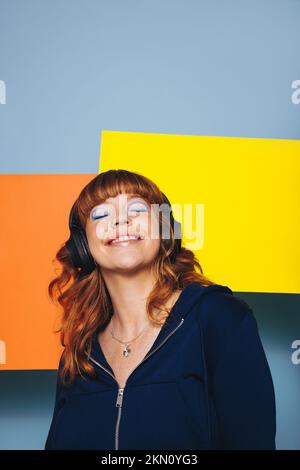 Lächelnde Frau, die Musik hörte, während sie vor zwei Werbetafeln in einem Studio stand. Glückliche junge Frau genießt eine Audio-Playlist über sie Stockfoto