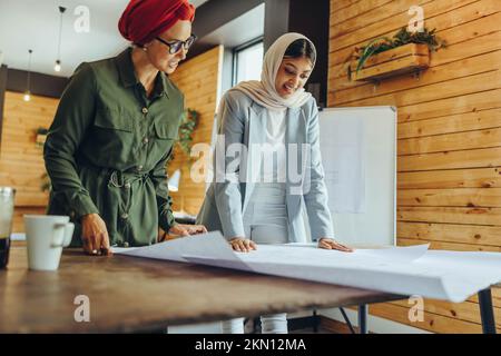 Kreative Architekten, die an Bauzeichnungen in einem modernen Büro arbeiten. Zwei muslimische Geschäftsfrauen planen ein neues innovatives Projekt. Zwei Designer Stockfoto