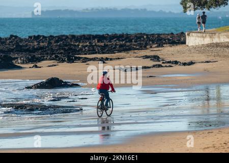 Ein Radfahrer in roter Jacke, der am Strand reitet. Unscharfe Leute, die in der Ferne laufen. Milford Beach, Auckland. Stockfoto
