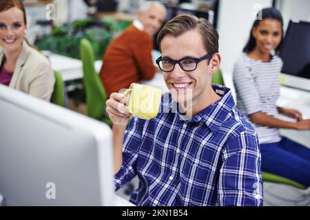 Ich habe das beste Team hinter mir. Porträt eines Mannes, der Kaffee trinkt, mit Kollegen, die im Hintergrund an Computern sitzen. Stockfoto