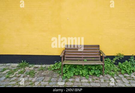 Ländliche Entspannung. Eine malerische Holzbank, die vor einer gelben Wand steht. Stockfoto