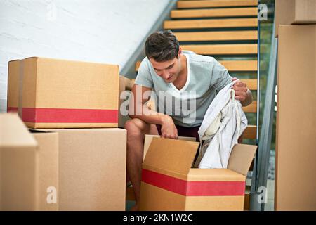 Nun zum Auspacken... ein junger Mann, der in seinem Haus sitzt, umgeben von verpackten Pappkartons. Stockfoto