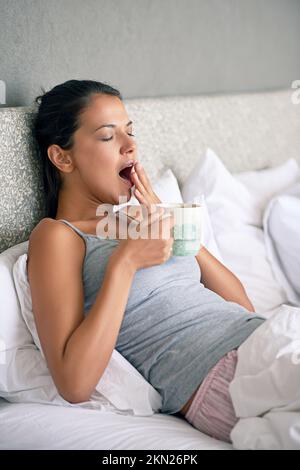 Es geht nichts über einen kleinen Kaffee, um einen aufzuwecken. Eine junge Frau, die Kaffee trinkt, während sie im Bett gähnt. Stockfoto