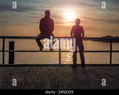 Ein paar Touristen. Eine Frau lehnt sich über das Geländer und sieht den Sonnenuntergang über dem Meer. Ein Mann sitzt auf einem Geländer und entspannt sich. Seebrücke, Rügen Stockfoto