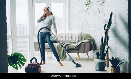 Die schöne junge Frau saugt zu Hause mit einem modernen Staubsauger auf den Boden und hört mit Kopfhörern Musik, tanzt und singt. Hausarbeit und Technikkonzept. Stockfoto