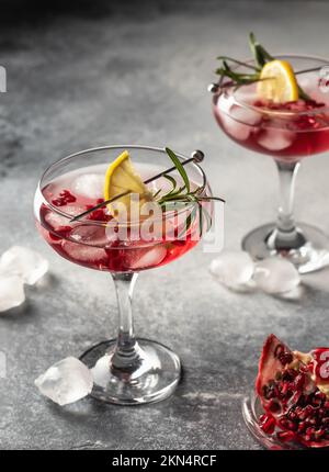 Zwei Gläser Granatapfel-Weihnachtscocktail mit Rosmarin und Zitrone auf grauem Hintergrund. Weihnachtsgetränke, Partykonzept, Menü Stockfoto