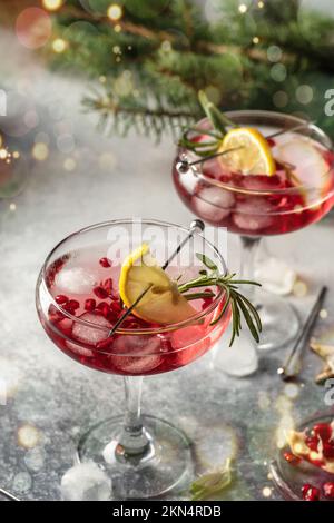 Granatäpfel-Eiscocktail mit Rosmarin und Zitrone auf grauem Hintergrund mit Bokeh-Lichtern und Tannenzweigen. Weihnachtsgetränk-Party-Konzept Stockfoto