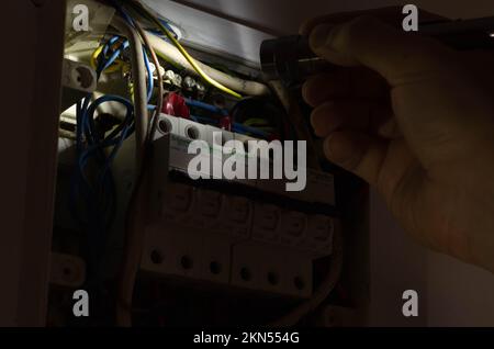 Blackout-Konzept. Die Hand einer Person in völliger Dunkelheit hält eine Taschenlampe in der Hand, um einen Sicherungskasten im Haus während eines Stromausfalls zu untersuchen. Stockfoto