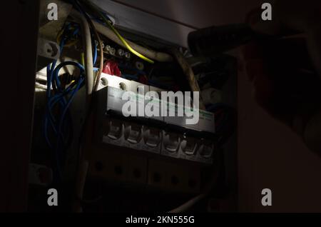 Blackout-Konzept. Die Hand einer Person in völliger Dunkelheit hält eine Taschenlampe in der Hand, um einen Sicherungskasten im Haus während eines Stromausfalls zu untersuchen. Stockfoto