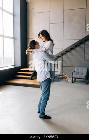 Glückliches, liebevolles Paar, das sich küsst, sich aufgeregt fühlt, in ein neues Zuhause zu ziehen, ein Mann, der eine Frau in den Händen hält, indem er beginnt, zusammen zu leben Stockfoto