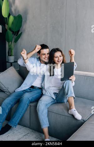 Aufgeregtes Paar, das Tablet und Quittungen überprüft, sitzt auf dem Sofa im Wohnzimmer zu Hause Stockfoto