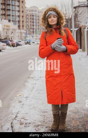 Mädchen mit Kaffee in warmer Jacke und Pelzhaube auf der Straße. Wintermode. Hübsche Frau in orangefarbener Kapuzenjacke in der Stadt. Urbaner Lebensstil Stockfoto