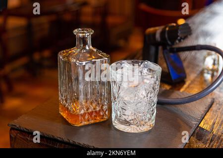 Whisky-Glaskaraffe mit Whisky-Likör und ein leeres Glas Whiskey mit einem Eis auf Schieferteller. Rauchmaschine im Hintergrund. Stockfoto