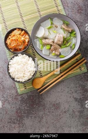 Muguk ist eine einfache Suppe mit koreanischem Rettich und Rindfleisch, serviert mit Reis und Kimchi als Nahaufnahme auf der Matte auf dem Tisch. Vertikale Draufsicht von oben Stockfoto