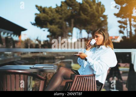 Elegante hispanische Frau, die Kaffee auf einer Terrasse in einer Umgebung mit Bäumen trinkt. Eine brünette Frau mit glattem Haar, die ein o trägt Stockfoto
