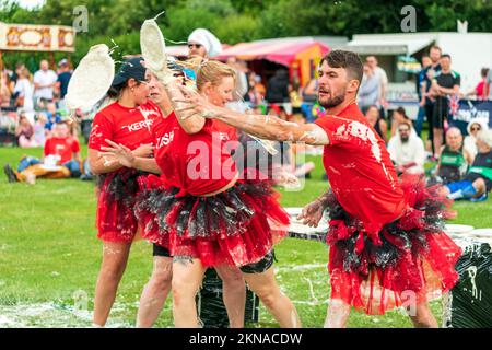 Ein gemischtes Team aus vier Personen in schicken Kleidern, teilweise mit Vanillesauce bespritzt, wirft Vanillesauce von einem Tisch während der World Custard Pie Championships. Stockfoto