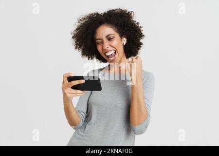 Junge hispanische Frau mit Kopfhörern, die online auf einem Handy spielt, isoliert vor blauem Hintergrund Stockfoto