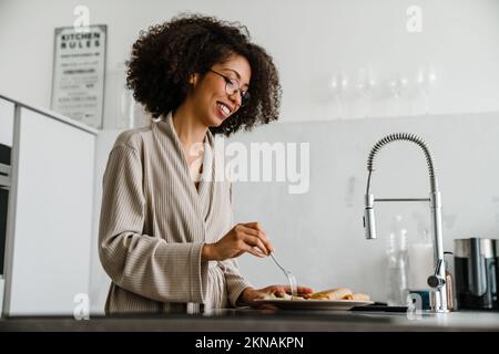 Eine schwarze Frau, die eine Brille trägt und lächelt, während sie zu Hause frühstückt Stockfoto