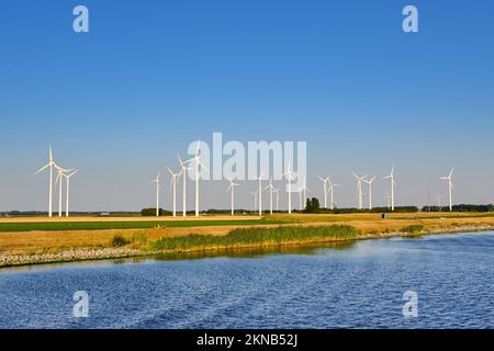 Hohe Windturbinen auf Ackerland entlang eines Kanals bei Abendlicht. Keine Menschen. Stockfoto