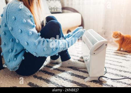 Heizelement im Winter zu Hause verwenden. Eine Frau, die ihre Hände mit einer Katze wärmt, die einen Pullover trägt. Heizperiode. Stockfoto